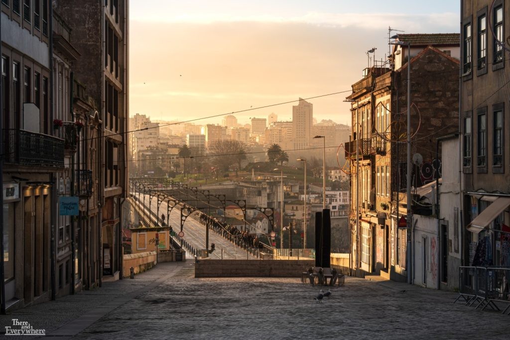 Early morning light shining down a cobblestone street toward the Luís I Bridge in Porto, Portugal, with a crowd waiting for the light rail.