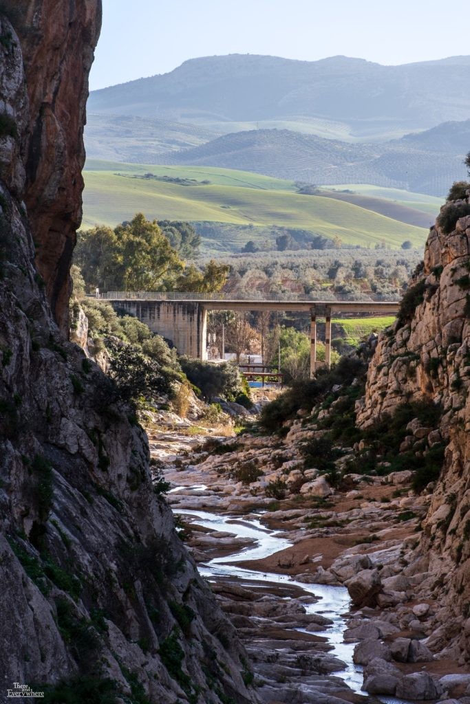 A rocky canyon view of Río de la Venta in Málaga, Spain, with a concrete bridge crossing the winding riverbed between steep cliffs.