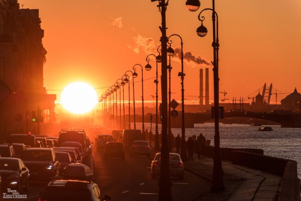 Cars driving along Admiralty Embankment at sunset in St Petersburg, Russia, silhouetted against a bright sun with streetlights and factory smoke in the distance.
